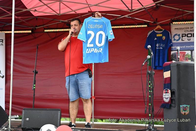 mężczyzna ubrany w krótkie spodenki stoi na scenie trzymając niebieską koszulkę z liczbą 20 i nazwiskiem Zieliński